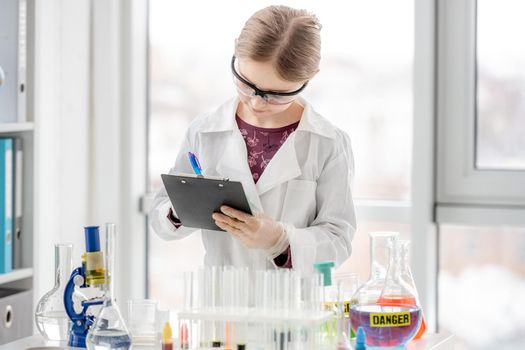 Girl during scientific chemistry experiment making notes close to table with tubes. Schoolgirl with chemical equipment on school lesson