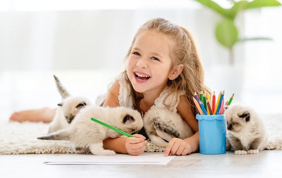 Child girl painting with ragdoll kittens on the floor and smiling. Little female person drawing with colorful pencils and kitty pets close to her at home