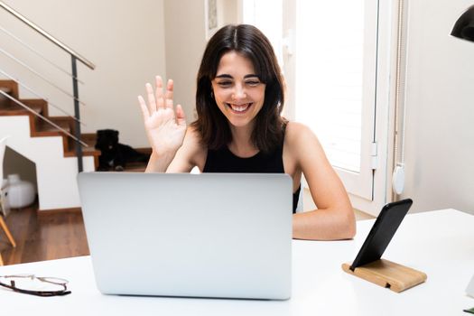 Happy and smiling young woman waving hello on video call using laptop at home. Technology concept.