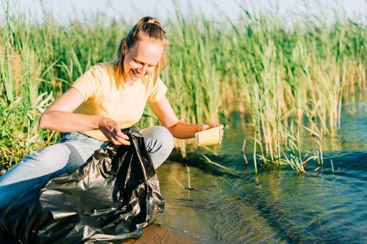 Young female volunteer satisfied with picking up trash, a plastic bottles and coffee cups, clean up beach with a lake. Woman collecting garbage in bag. Environmental ecology pollution concept.