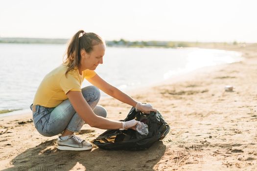 Young female volunteer satisfied with picking up trash, a plastic bottles and coffee cups, clean up beach with a sea. Woman collecting garbage. Environmental ecology pollution concept. Earth Day.