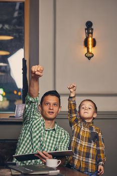 Young father and his stylish little son in the barbershop in the waiting room. They are waiting for the master and having a good time. They choosing haircut from the mahazine