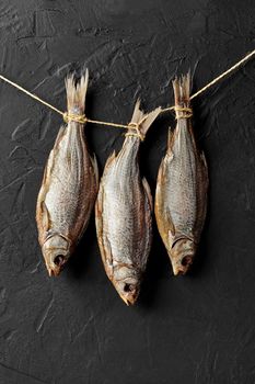 Three dried salted roach fish hanging by tails on rope on black wall