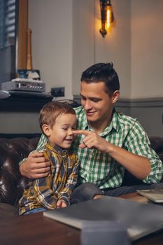 Young father and his stylish little son in the barbershop in the waiting room. They are waiting for the master, reading the fashion magazine and having a good time. Father hugs his son