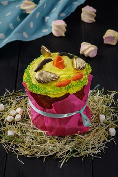 Funny Easter cake wrapped in pink paper cover, decorated with yellow whipped egg whites in shape of cute chicken standing on hay on black wooden table with marshmallows. Fancy festive baked goods