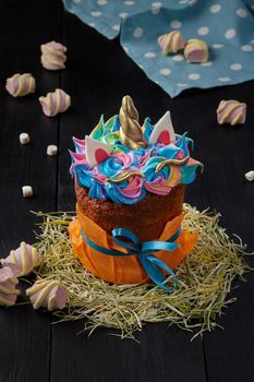 Fancy Easter cake in orange paper wrapping decorated with colorful whipped egg whites in shape of unicorn with golden horn, standing on hay on black wooden table with marshmallows. Festive baked goods