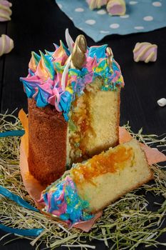 Sliced Easter cake from soft tender dough with fruit filling decorated with colorful whipped egg whites in shape of unicorn with golden horn in orange paper wrapping on hay on black wooden background