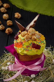 Easter cake in colored paper wrapping decorated with yellow whipped egg whites, caramel popcorn, gummy bears and candy bar slices standing on hay on black wooden table. Fancy festive baked goods