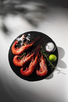 Whole raw giant red prawns on round black slate serving board with condiments, lime, clove of garlic and fresh rosemary on gray background with shadow. Seafood appetizer. Flat lay with copyspace