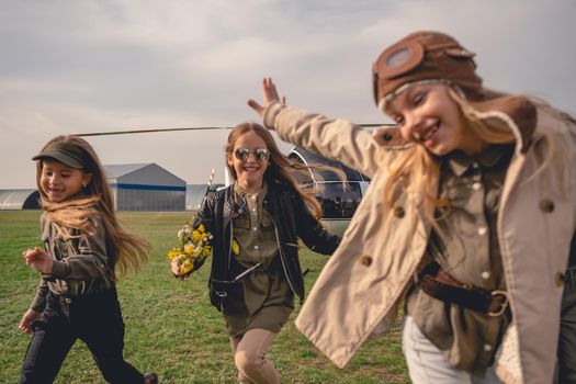 Group of cheerful carefree tween girls enjoying time together, running on flying field of aeroclub on background of standing helicopter on cloudy day in spring