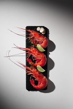 Top view of three whole cooked royal prawns on black slate board with lime, garlic and fresh herbs on grey background with shadow. Seafood delicacies