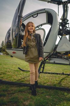 Smiling cute brown-haired preteen girl standing near open door of white small helicopter on flying club field on cloudy spring day. Full length portrait