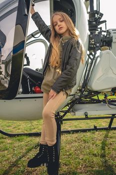 Portrait of dreamy tween girl standing on steps of white modern helicopter outdoors, holding by assist handle of cockpit