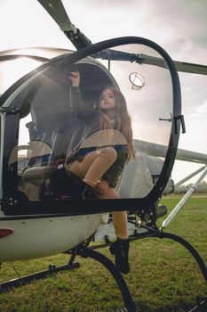 Preteen girl sitting in cockpit of landed modern helicopter in pilots seat behind glass of open door in rays of autumn sun breaking through clouds