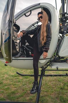 Confident brown-haired tween girl wearing black leather jacket and sunglasses standing on footboard of open helicopter landed at airfield