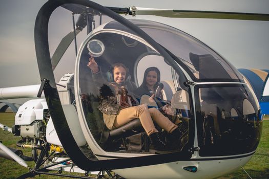 Cheerful tween girl wearing aviator headphones sitting on co-pilot seat in helicopter cockpit with her friend, pointing to sky