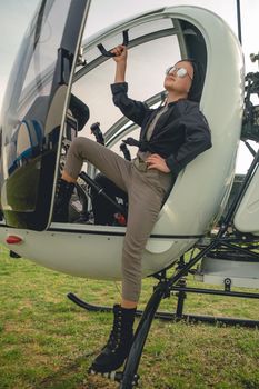 Stylish preteen girl in mirrored sunglasses looking up to sky while standing on steps of open helicopter landed on green flying field in springtime