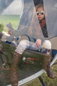 View through glass of helicopter cockpit of confident preteen girl in mirrored sunglasses sitting on pilots seat, holding control handle on cloudy autumn day