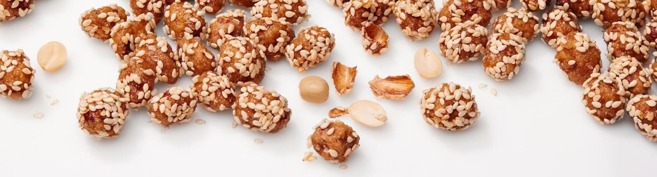 Closeup of delicious candied peanuts in honey and sugar glaze, sprinkled with sesame scattered on white surface. Concept of sweet cracker nuts as snacks. Horizontal image of natural food background