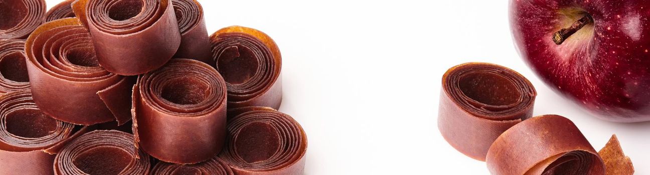 Closeup of rolls of fruit leather stacked on white surface with fresh ripe red apple. Concept of popular fruit sweets and organic snacks. Natural food background with copyspace