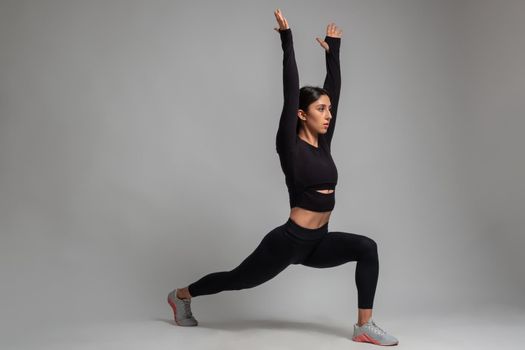 Concentrated fit brunette in black sportswear performing leg and glute workout, doing bodyweight forward lunges with arms stretched straight upwards in studio on grey background. Fitness concept
