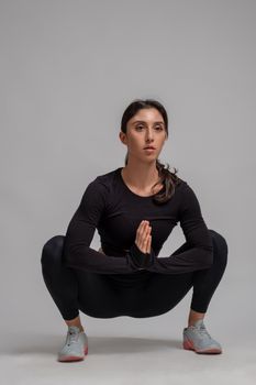 Focused athletic girl in black sportswear doing stretching squats before workout on grey background. Fitness and body conscious concept