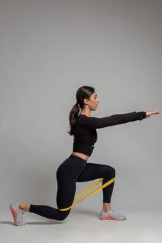 Sporty girl in black activewear working out leg and glute muscles, performing lunges with resistance band on grey background. Physical activity and fitness concept