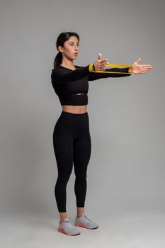 Athletic girl in black sportswear doing exercise to sculpt and tone arms, stretching resistance elastic band on outstretched arms on grey background. Sport and fitness concept