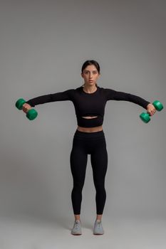 Sporty girl in black activewear working out shoulders muscles in studio on grey background, performing dumbbell lateral raise. Physical activity and fitness concept