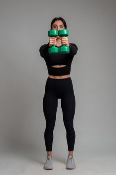 Sporty girl in black activewear working out shoulders muscles in studio on grey background, performing exercises with dumbbells. Physical activity and fitness concept