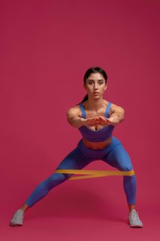 Sporty girl working out muscles of legs and glutes, doing lateral lunges with resistance loop band in studio on maroon background. Fitness and sports motivation concept