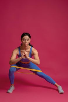 Athletic girl working out in studio interior on maroon background, doing lateral lunge to right with resistance loop band. Leg and glute workout. Active lifestyle and fitness concept