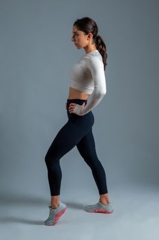 Side view of confident fit girl wearing sports top and leggings standing with arms akimbo, posing during workout. Studio full length portrait on grey background
