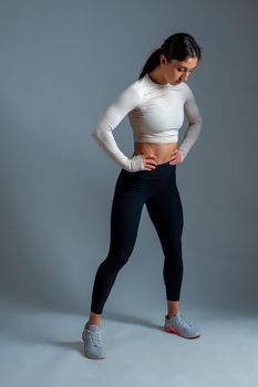 Full length portrait of young fit brunette wearing sports top and leggings standing with arms akimbo, posing during workout in studio on grey wall background