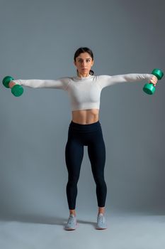 Athletic girl in activewear working out shoulders muscles in studio on grey background, performing dumbbell lateral raise. Physical activity and fitness concept