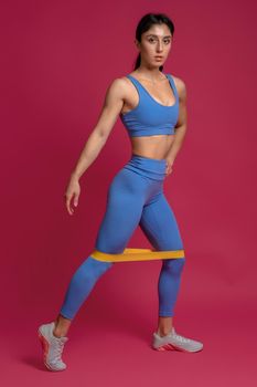 Portrait of focused sporty girl in blue activewear doing lower body exercises with resistance band on maroon background. Fitness and strength training concept