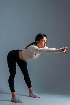 Focused athletic girl in sportswear doing pre-workout stretch, bending body forward with outstretched arms on grey background. Fitness and body conscious concept