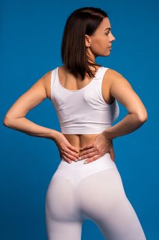 Back view of fit dark-haired woman in white sports top and leggings standing against blue background. Fitness and self-improvement concept