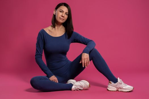 Relaxed young brunette wearing yoga jumpsuit sitting on floor on maroon background, resting after workout and looking at camera