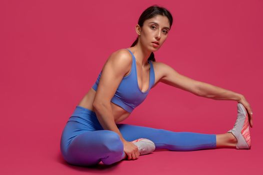 Young sportswoman warming up before intense workout, doing stretching exercises sitting on floor and looking confidently at camera. Studio portrait on maroon background