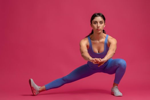 Young fit woman performing bodyweight side lunges during leg and glute workout, looking confidently at camera. Studio full length portrait on maroon background