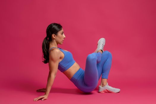 Side view of young athletic brunette performing stretching workout to improve muscle elasticity and achieve comfortable muscle tone. Studio shot against maroon background