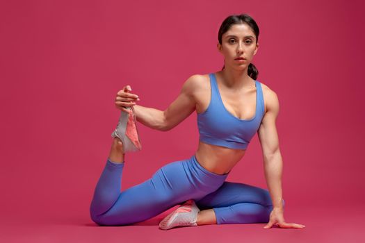 Flexible young woman wearing blue sportswear exercising on maroon background, doing stretching workout. Fitness, bodyweight training, active lifestyle concept