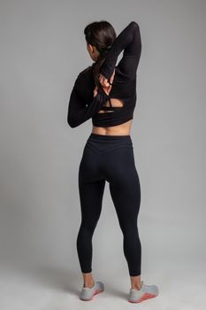 Rear view of sporty flexible girl doing stretching workout, standing with fingers clasped behind her back. Sportswoman training in studio on grey background