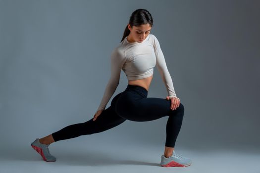 Young athletic brunette performing stretching lunges with body twists to improve muscle elasticity and achieve comfortable muscle tone. Fitness concept. Studio shot against grey background