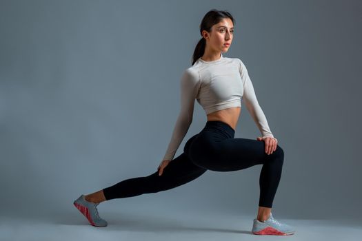 Confident young sportswoman warming up before intense workout, doing stretching lunges with twisting of body. Full length studio portrait on grey background