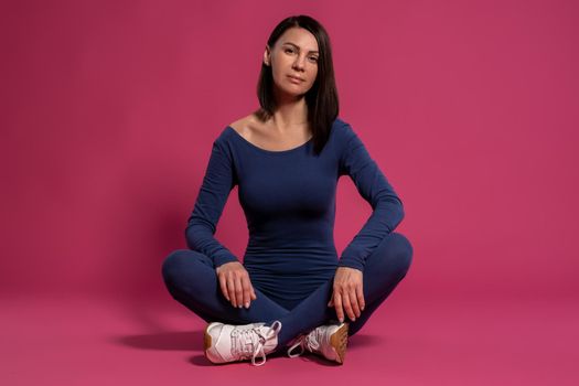 Calm slim woman in fitness jumpsuit sitting cross-legged on floor, relaxing after training. Studio portrait on maroon background