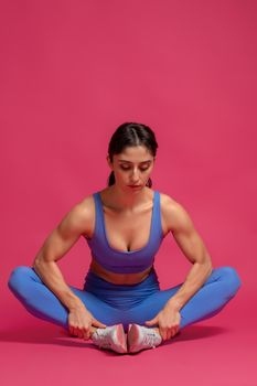 Focused female athlete stretching before training, preparing muscles of body for intense workout, sitting on floor on maroon background. Health and sport concept