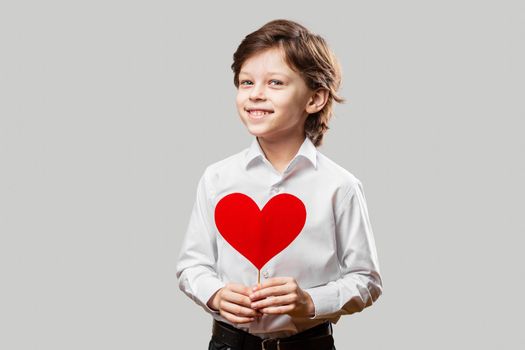 White boy wearing white shirt holding a red heart and smiling. Celebrating St. Valentine’s Day or Mother’s Day.