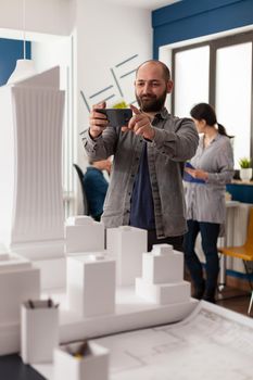 Smiling design architect holding smartphone to photograph desk with architectural model of skyscraper and buildings. Bearded engineer happy with final white foam maquette posting on social media.